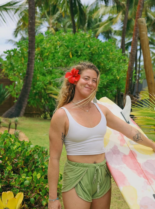 Kyla Peterson with a hibiscus flower in her hair and surfboard under her arm in a tropical setting